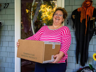 A woman in a pink shirt smiles on a porch holding a box of vegetables to mail