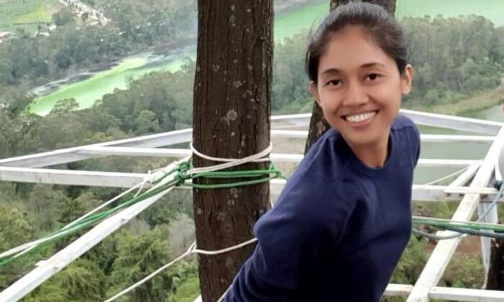 Nety Riana Sari stands beside a tree overlooking a forest and smiles at the camera