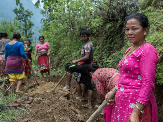 A group of people standing on a mountainside digging with a woman in all pink standing in the forefront looking at the camera