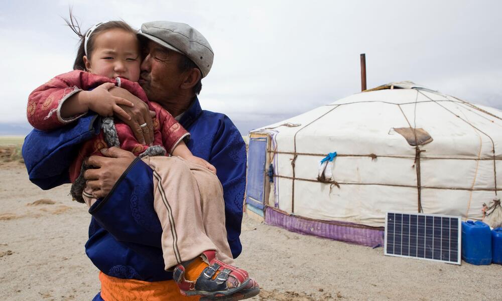 A father holds his young daughter in his arms and kisses her on the cheek in front of a yurt