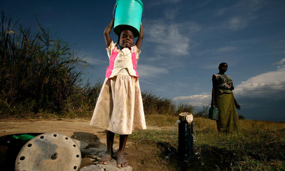 A girl in a white dress holds a green bucket above her head and smiles at the camera