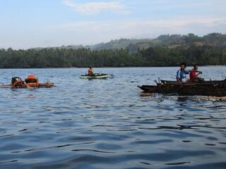 men on small wooden boats paddle across calm blue water with a coastline of trees in the background