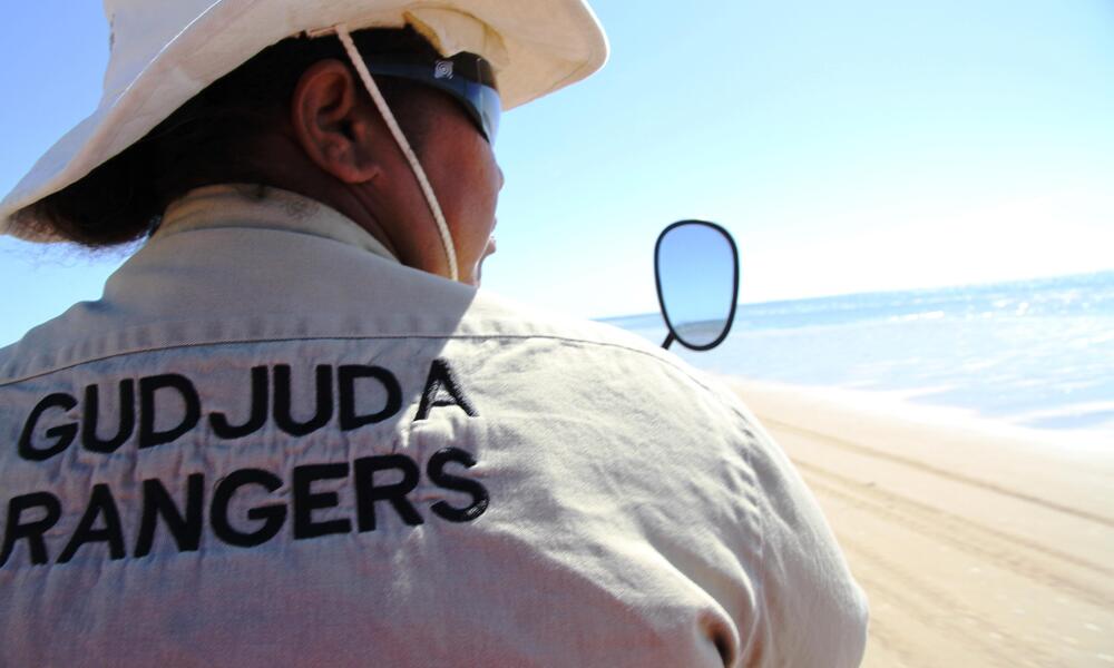 View of a person's back against the beach. Wearing a khaki hat and jacket that says "Gudjuda Rangers"